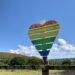 The image shows a landscape with blue sky and a heart-shaped wooden sign in rainbow colours.