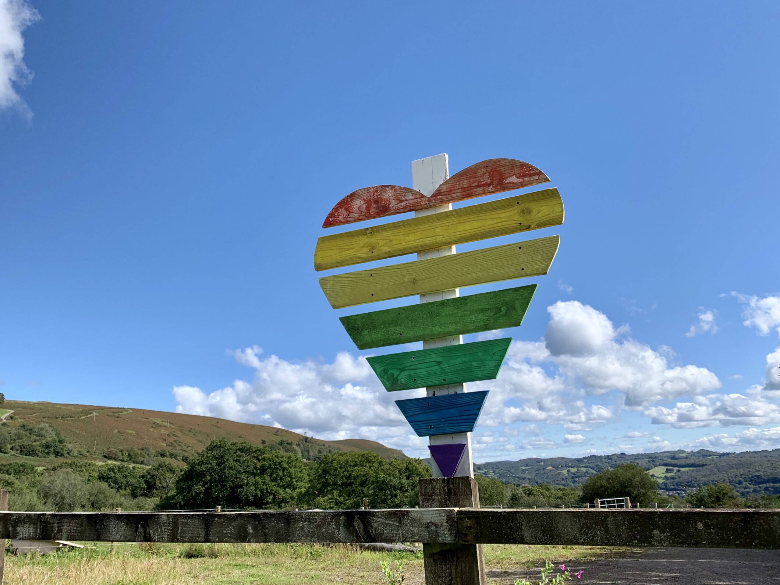 The image shows a landscape with blue sky and a heart-shaped wooden sign in rainbow colours.